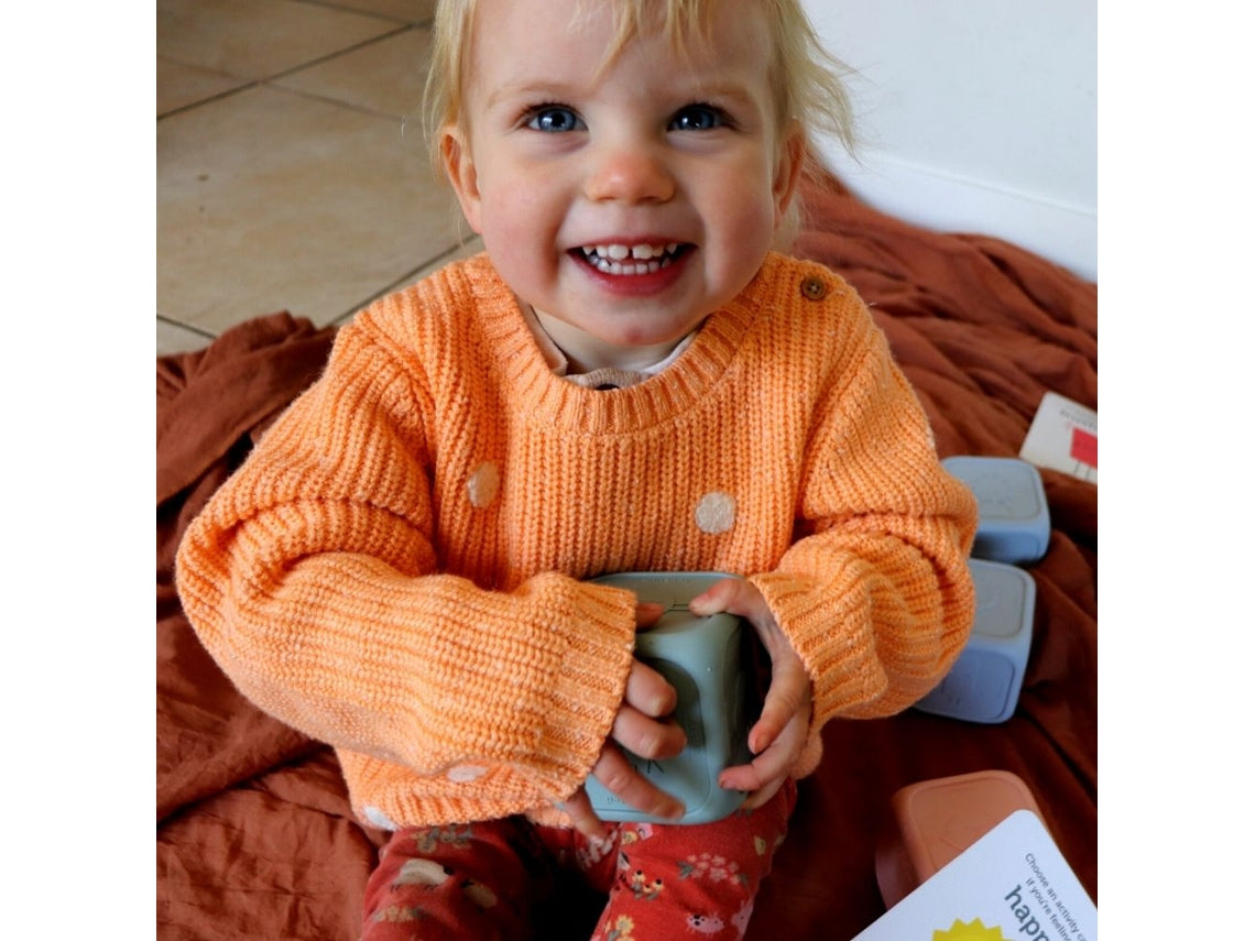 Criança pequena com camisola laranja sorri enquanto segura o cubo de sentimentos Sage Mint nas mãos.