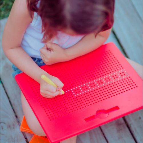Criança pequena, vista de cima, a utilizar a caneta amarela para desenhar pontos luminosos no quadro magnético vermelho BRAINTOYS IMApad.