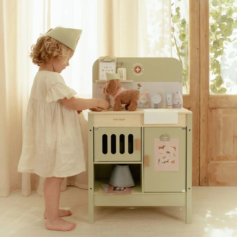Criança pequena de pé junto à estação veterinária de madeira, a interagir com um peluche de cão no lavatório da clínica.