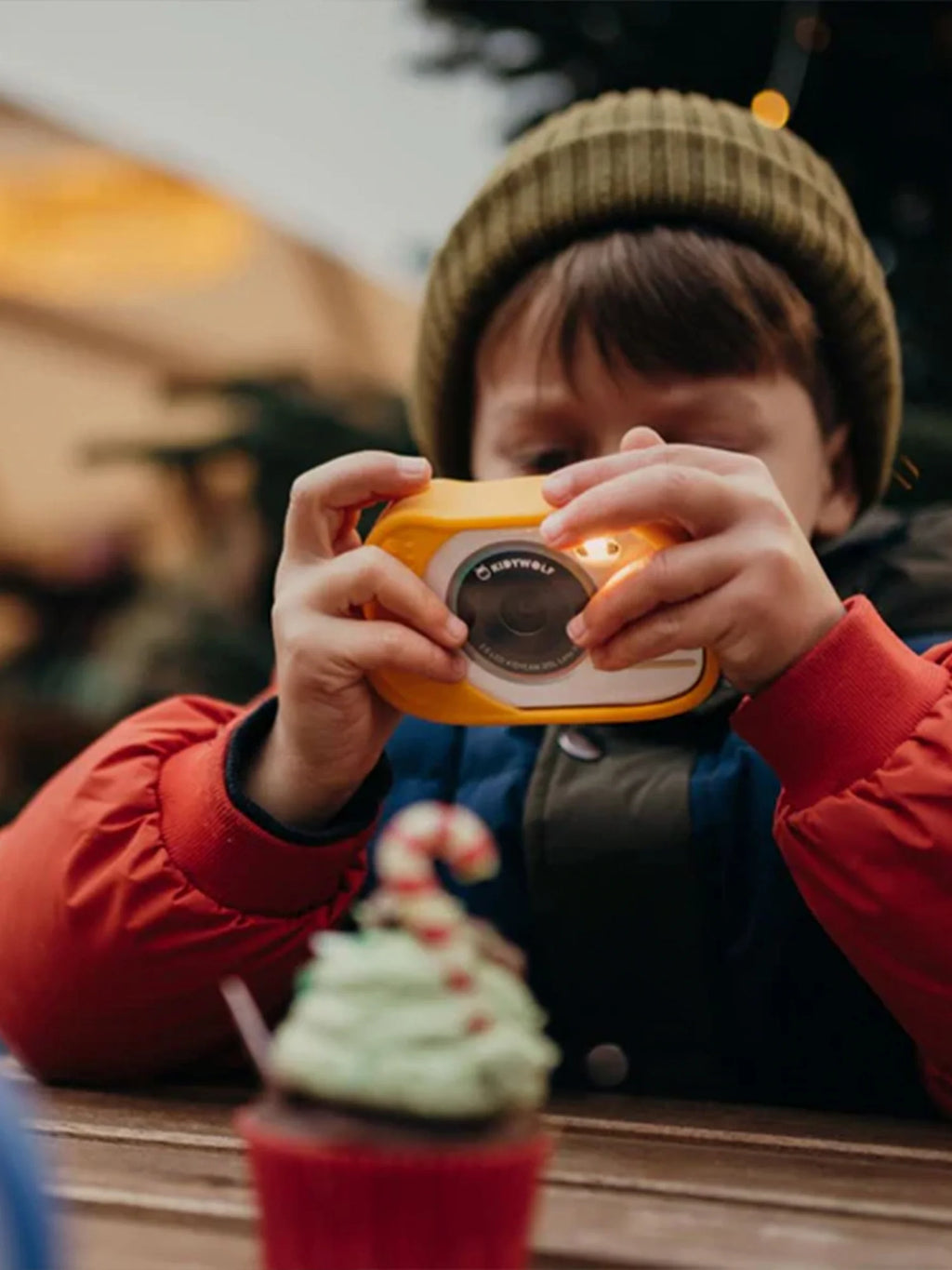 Criança com gorro a usar a máquina fotográfica Kidywolf amarela para tirar uma foto a um cupcake decorado, em ambiente exterior.