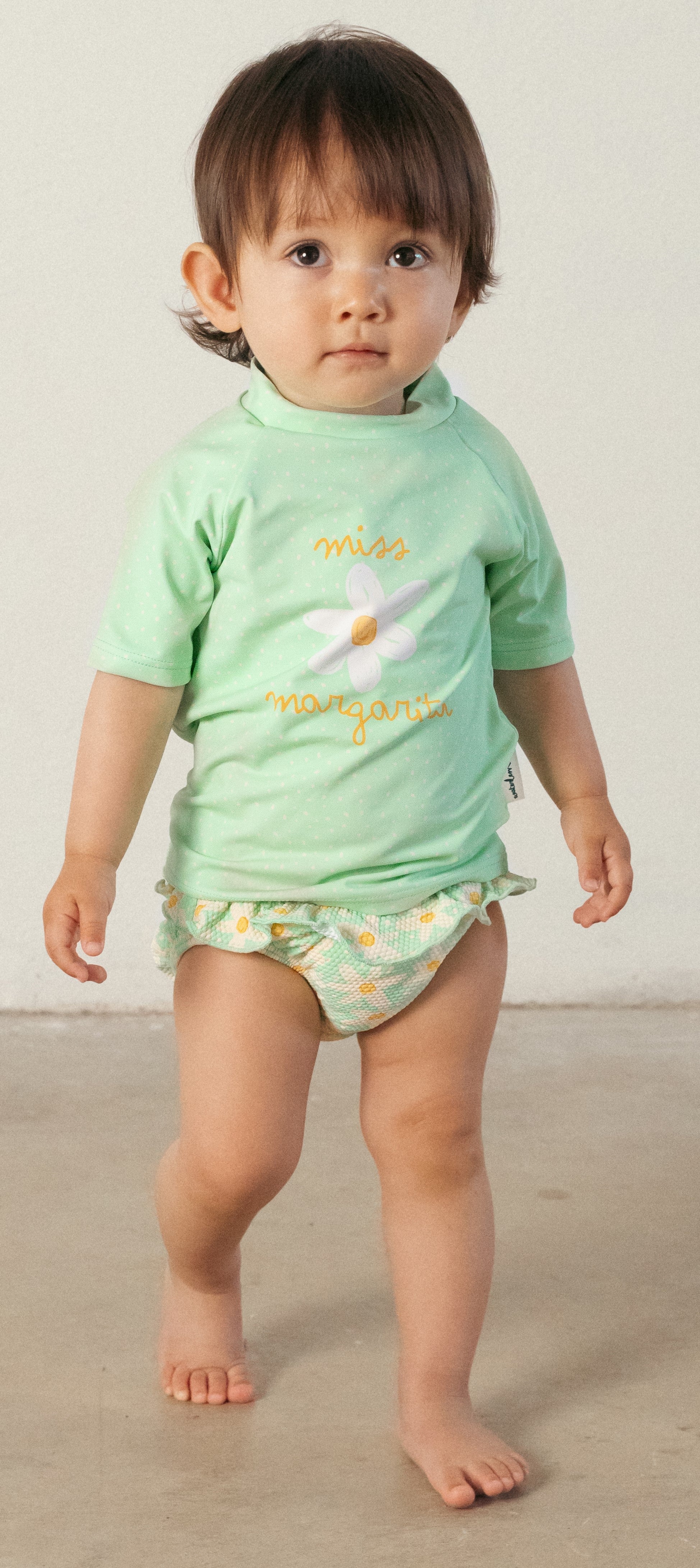 Criança pequena, de pé, a usar a fralda de praia Waterlemon verde com margaridas e uma t-shirt de proteção solar verde.