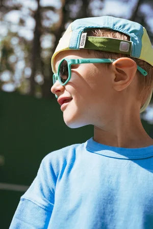 Criança sorridente a usar óculos de sol Kietla Eyzz turquesa e um boné, olhando para o lado, com um fundo verde desfocado.