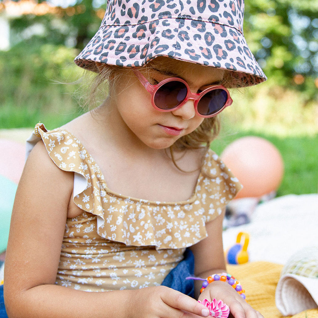 Criança ao ar livre a usar óculos de sol redondos rosa claro, chapéu de padrão leopardo e fato de banho floral.