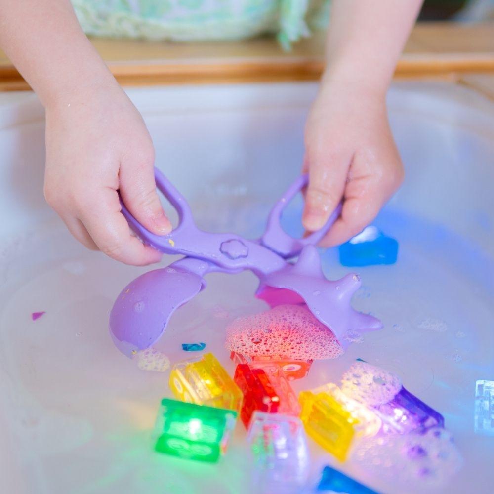Criança a usar as pinças GloPals para apanhar objetos numa mesa sensorial com água e espuma, desenvolvendo a motricidade fina.