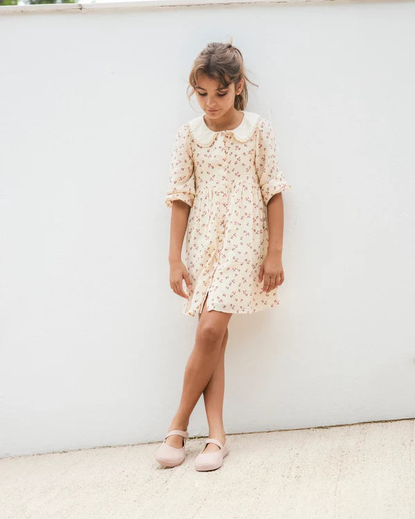 Criança em vestido floral com calçado barefoot Muris Sienna, encostada a uma parede branca.