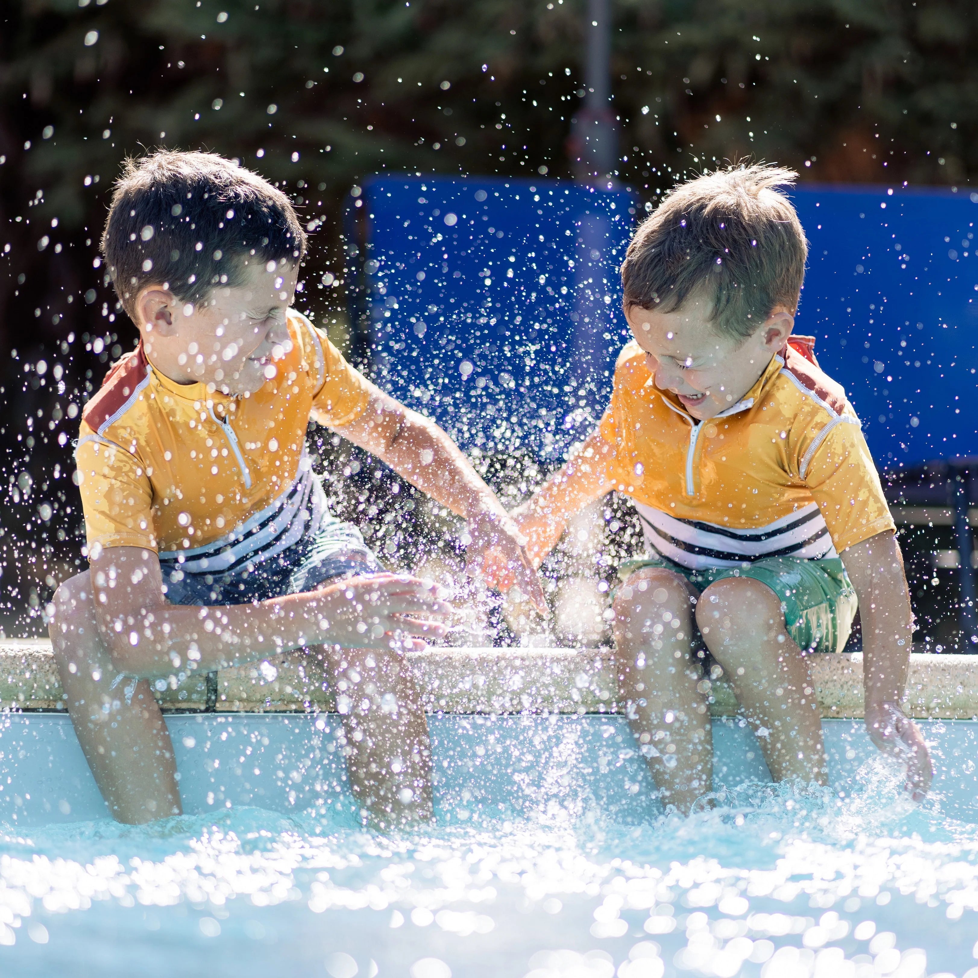 Duas crianças sorridentes a brincar na água, salpicando com os pés, ambas a usar a t-shirt de segurança Floatee amarela.
