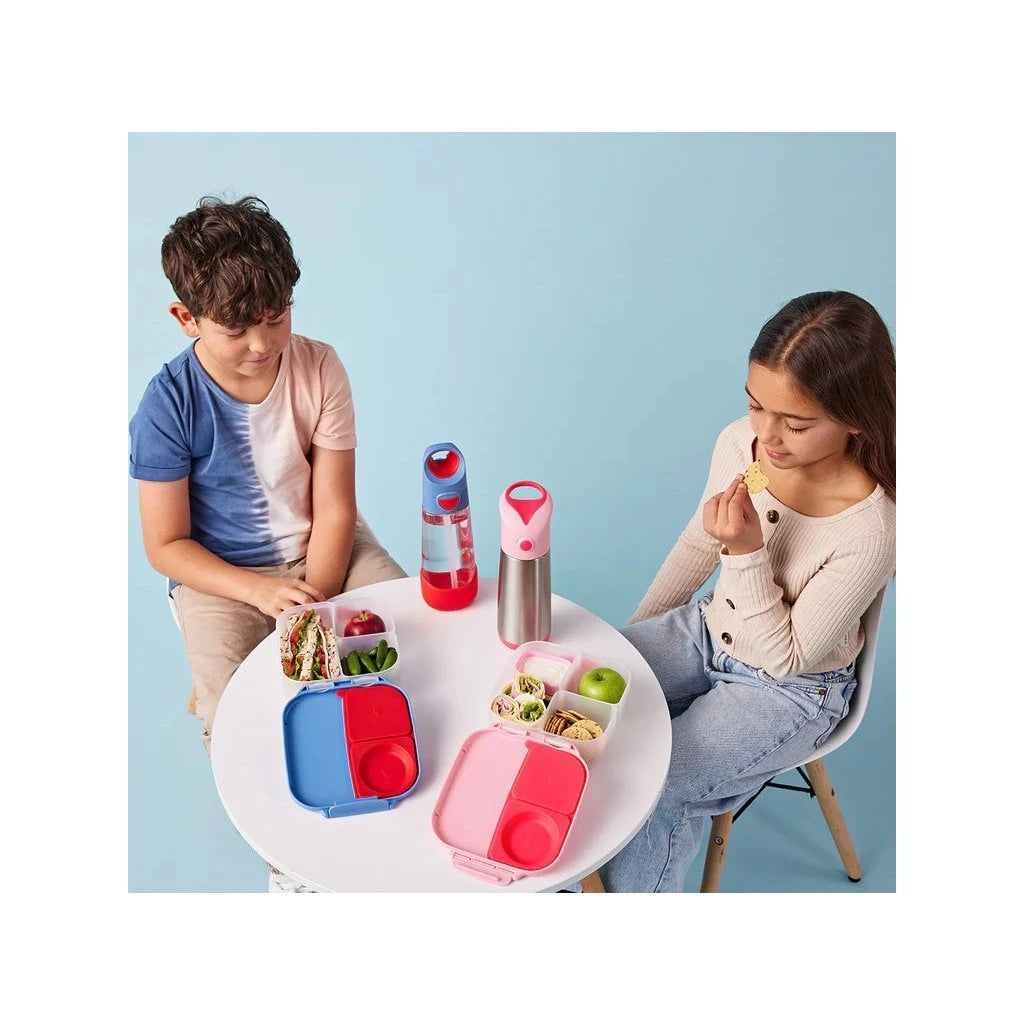 Duas crianças sentadas a uma mesa branca, cada uma com o seu recipiente de almoço (um rosa e um azul) aberto, a comer.