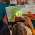Crianças a lerem um livro da Glo Pals, utilizando a lupa mágica para iluminar uma página com a imagem de um dinossauro verde.