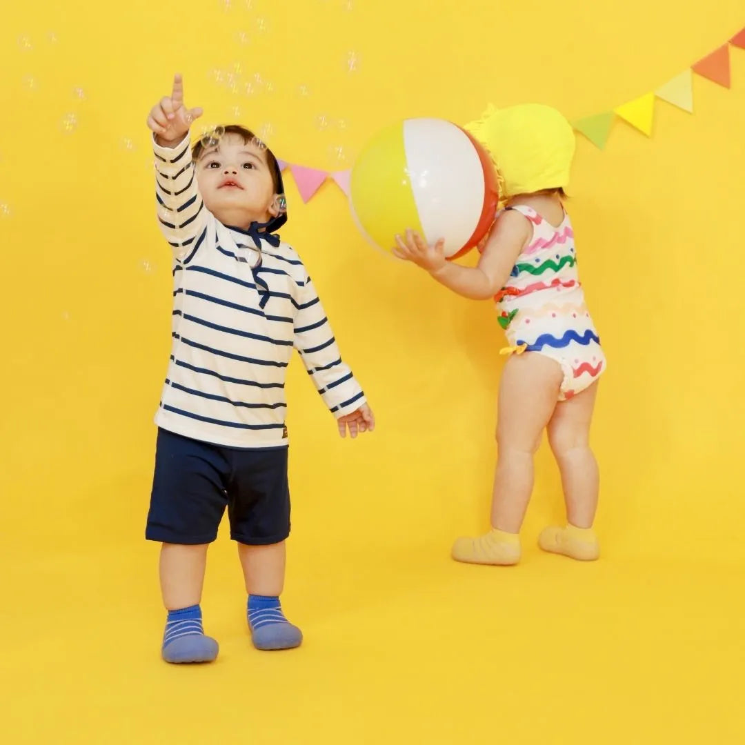  Duas crianças a brincar com uma bola num cenário amarelo vibrante, uma delas a usar o calçado barefoot Attipas See Through Yellow.