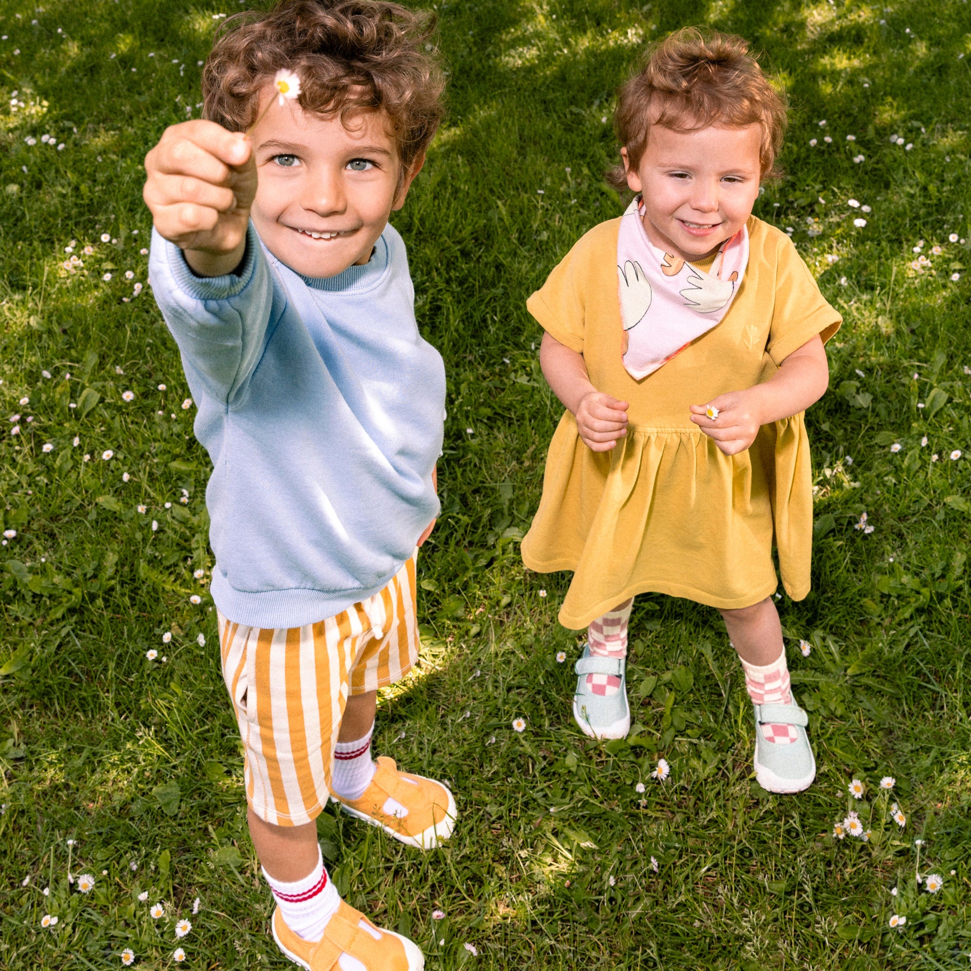 Duas crianças a brincar na relva com sandálias barefoot Victoria; uma criança usa um conjunto azul e amarelo, a outra um vestido amarelo, ambas sorridentes.