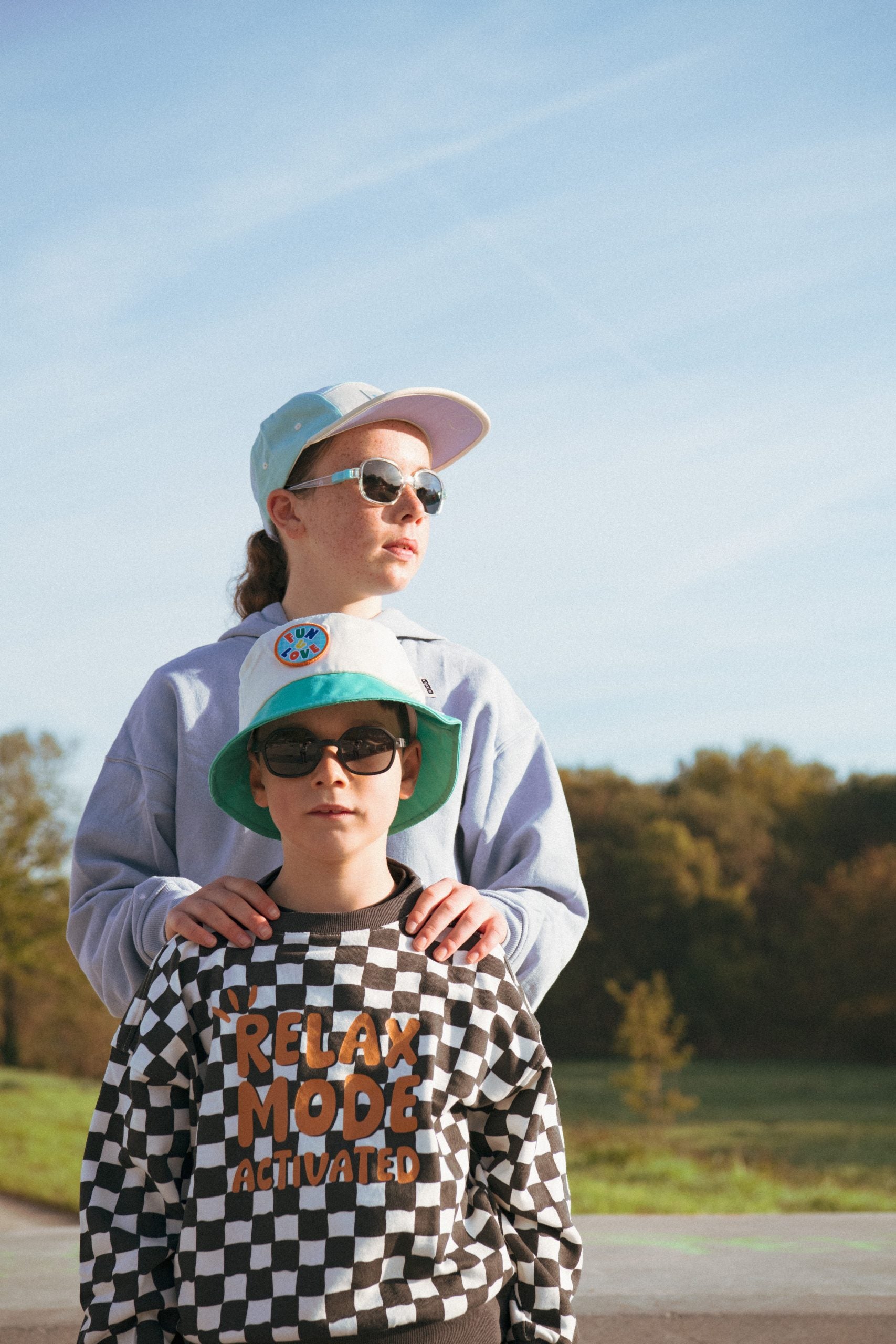Duas crianças ao ar livre, uma mais velha a usar óculos de sol e um boné, e um rapaz mais novo à frente com óculos de sol e um chapéu bucket.