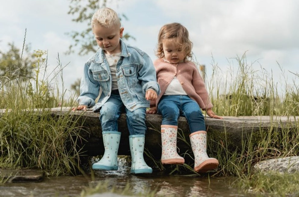 Duas crianças sentadas numa pedra junto à água, uma com galochas Little Dutch azuis e outra com galochas florais, preparadas para brincar na chuva.