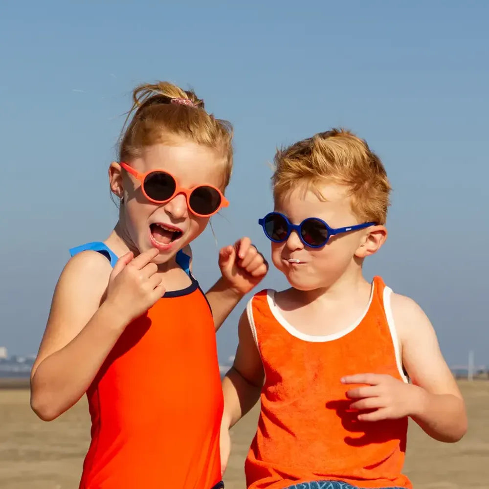 Duas crianças, um rapaz e uma rapariga, na praia ao sol, ambos a usar óculos de sol redondos (laranja e azul) e a divertirem-se.