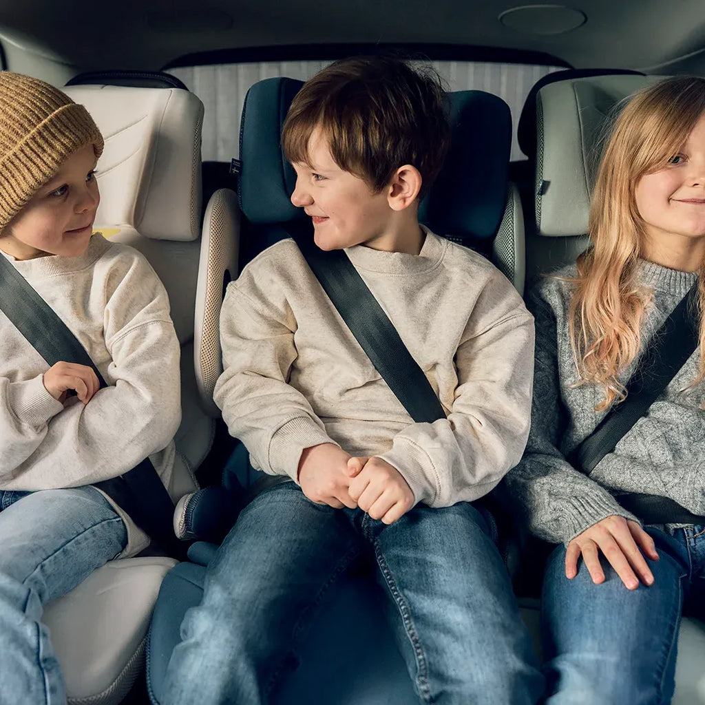 Três crianças sorridentes sentadas no banco traseiro de um carro, duas delas em cadeiras auto Axkid Up, a viajar a favor da marcha.