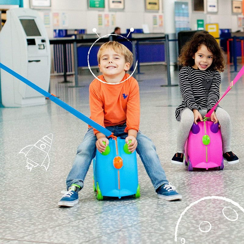 Duas crianças sorridentes em aeroporto, uma a andar numa mala Trunki azul e outra numa rosa, com ilustrações desenhadas de foguetes e planetas.