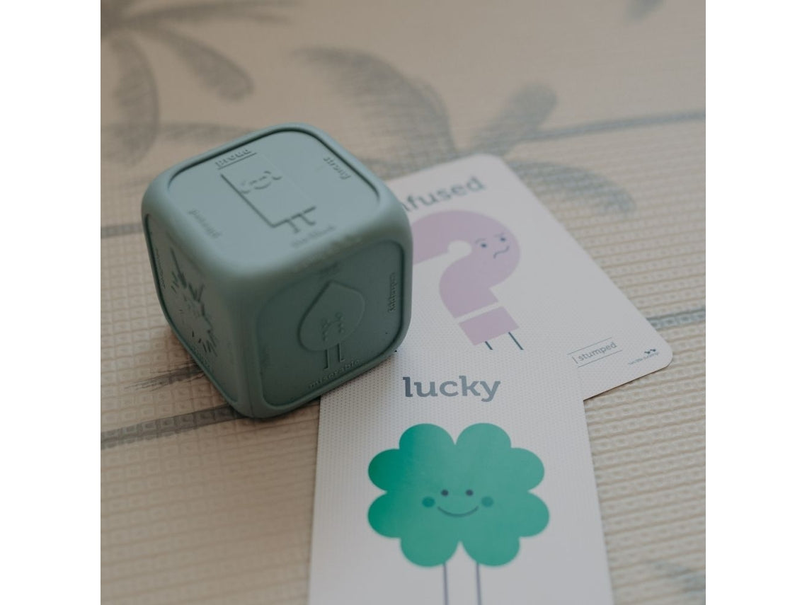 Cubo de silicone ao lado de cartões ilustrados com emoções, um deles mostrando um trevo verde para 'lucky'.