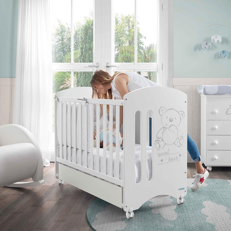 Mãe a colocar bebé no berço Micuna Sweet Bear 120x60 branco, com design de urso, em quarto infantil luminoso.