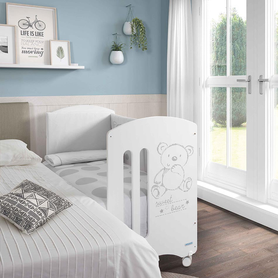 Berço Micuna Sweet Bear 120x60 branco, com design de urso, ao lado de cama de adulto, em modo colecho.