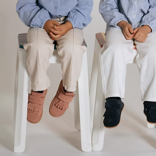 Duas crianças sentadas, uma com as Rutis Carneiras PiuPiu Rosa e outra com um modelo diferente.