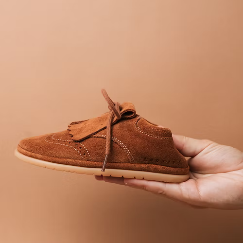 Mão a segurar uma sapatilha Rooties Carneiras Mémé Camel, realçando o seu design leve e compacto.