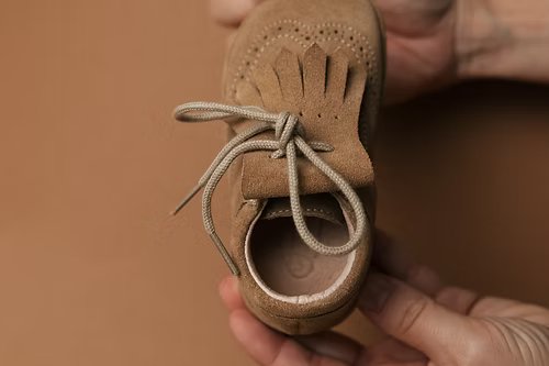 Detalhe superior da carneira Rooties Mémé Areia a ser segurada, mostrando os atacadores, a franja e a biqueira larga de estilo barefoot.