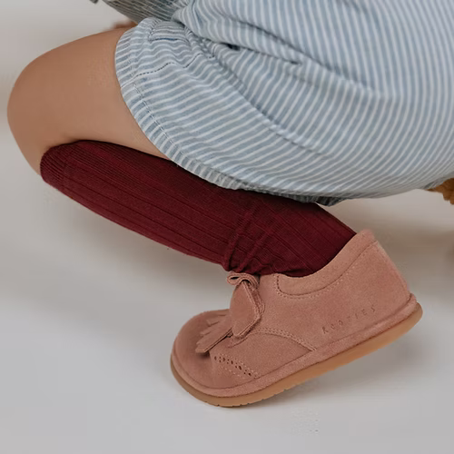 Criança agachada, detalhe do pé com a sapatilha Rutis Carneiras PiuPiu Rosa e meias bordeaux.