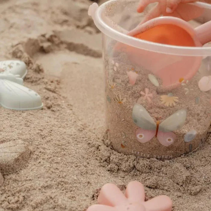 Detalhe do balde de praia transparente Little Dutch a ser enchido com areia, mostrando a textura do material e a decoração floral.