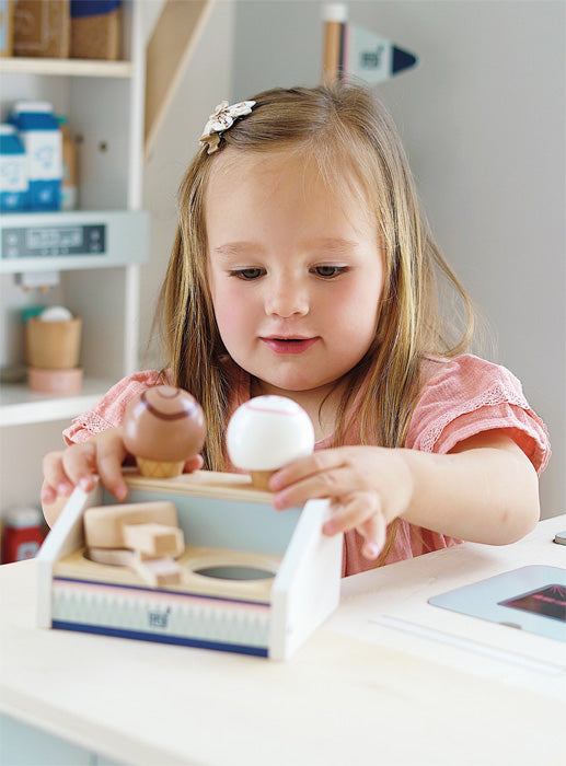 Uma menina brinca com a gelataria compacta de madeira small foot, servindo bolas de gelado com uma colher de madeira.