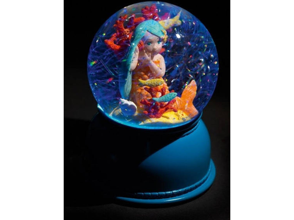 Globo de neve Djeco com luz mágica acesa, apresentando uma sereia de cabelo azul rodeada por corais e flocos de neve em movimento.