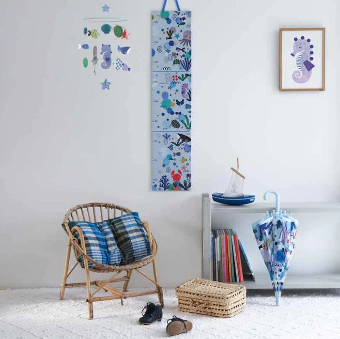 Quarto infantil decorado com a tabela de altura Djeco de vida marinha pendurada, móveis de vime, um móbile temático e um guarda-chuva azul.