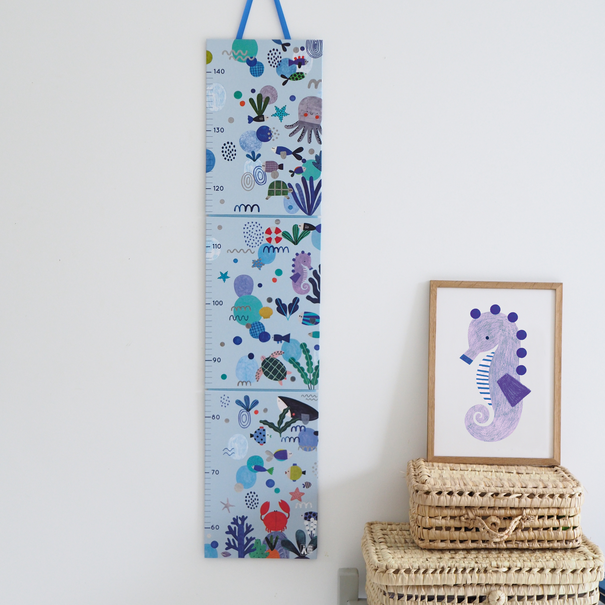 Tabela de altura Djeco "Aquatic Life" pendurada numa parede branca, decorada com ilustrações marinhas coloridas e graduada de 60 cm a 1,40 m. Ao lado, cestos de vime e um quadro com um cavalo-marinho.