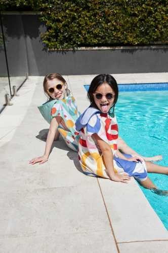 Duas meninas a divertirem-se à beira da piscina, sentadas e a mostrar os ponchos absorventes Dock and Bay com estampas abstratas e coloridas.