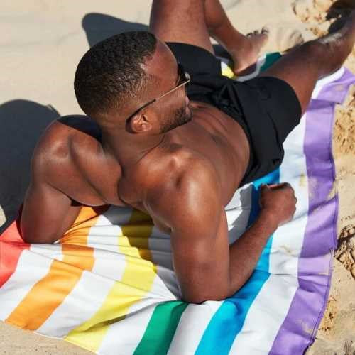 Homem bronzeado deitado de bruços sobre uma toalha de praia Dock and Bay com riscas de arco-íris, a apanhar sol na areia.