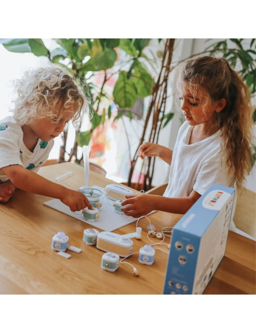 Duas crianças a brincar e a montar circuitos com o kit KIDYELEC na mesa, com a caixa por perto.