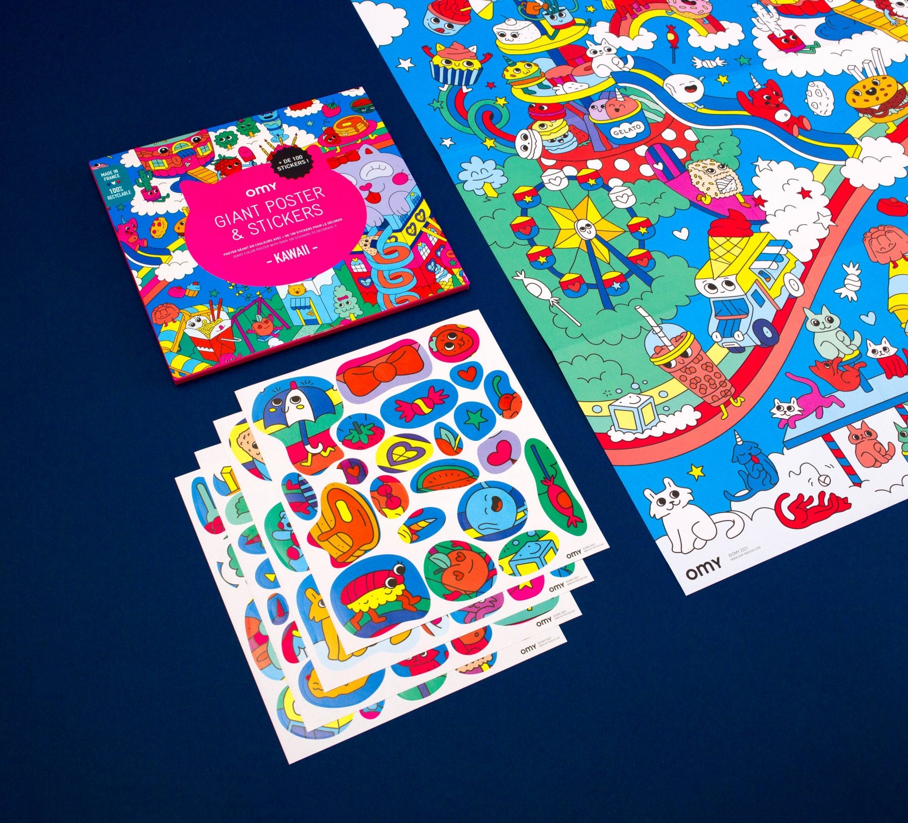 Vista aérea do conteúdo do OMY Poster Autocolantes XL - Kawaii, mostrando o poster desdobrado com ilustrações de um parque de diversões colorido e várias folhas de autocolantes com formas e personagens diversas.