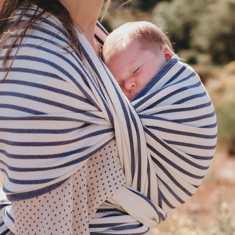 Mãe a carregar um bebé recém-nascido no Pano Tecido Neobulle T7 Malo, listrado em azul e branco, ao ar livre.