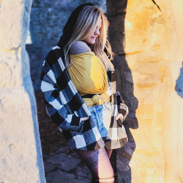 Mulher vestida com uma camisa de flanela xadrez preta e branca, calções de ganga e meias compridas, usando um pano elástico amarelo Hoppediz Curry para carregar um bebé. A mulher está num ambiente ao ar livre, entre duas paredes de pedra iluminadas pelo sol.