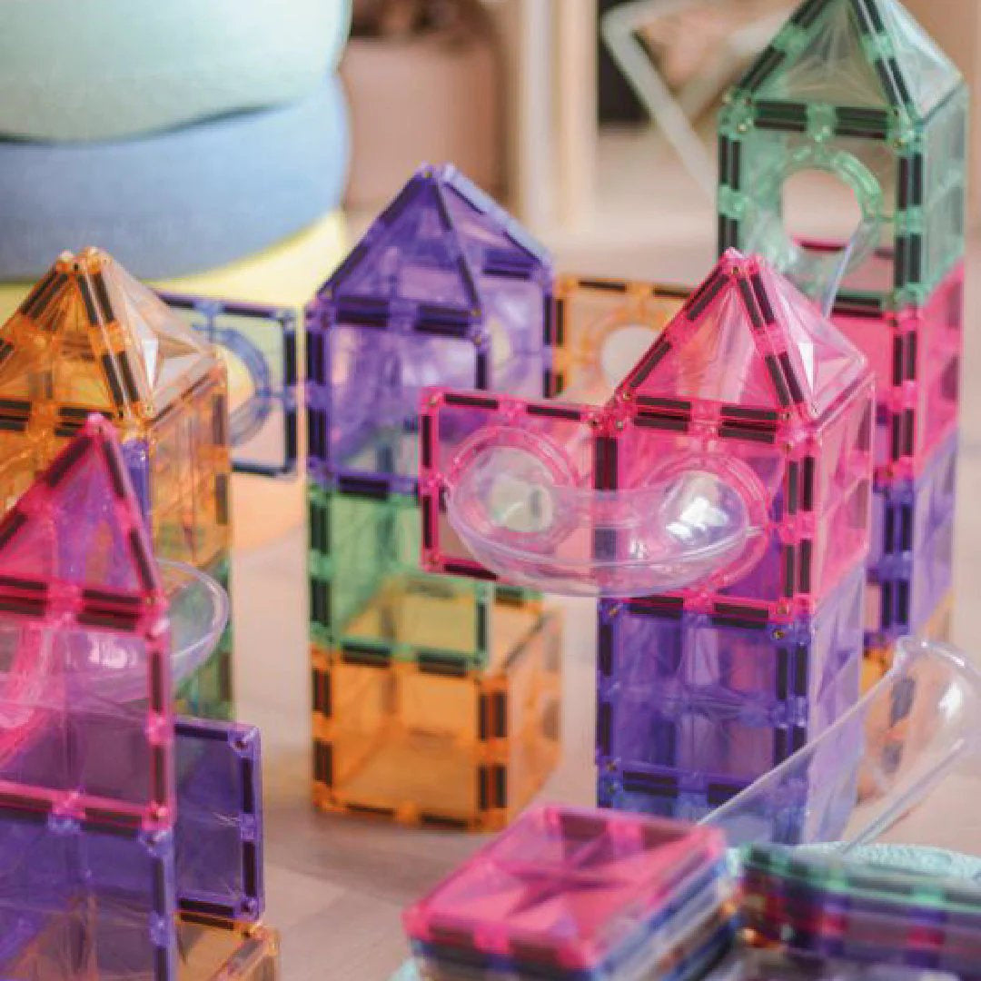 Construção de casas e torres com blocos magnéticos transparentes em tons pastel de rosa, laranja, roxo e verde, com tubos de pista de bola transparentes integrados.