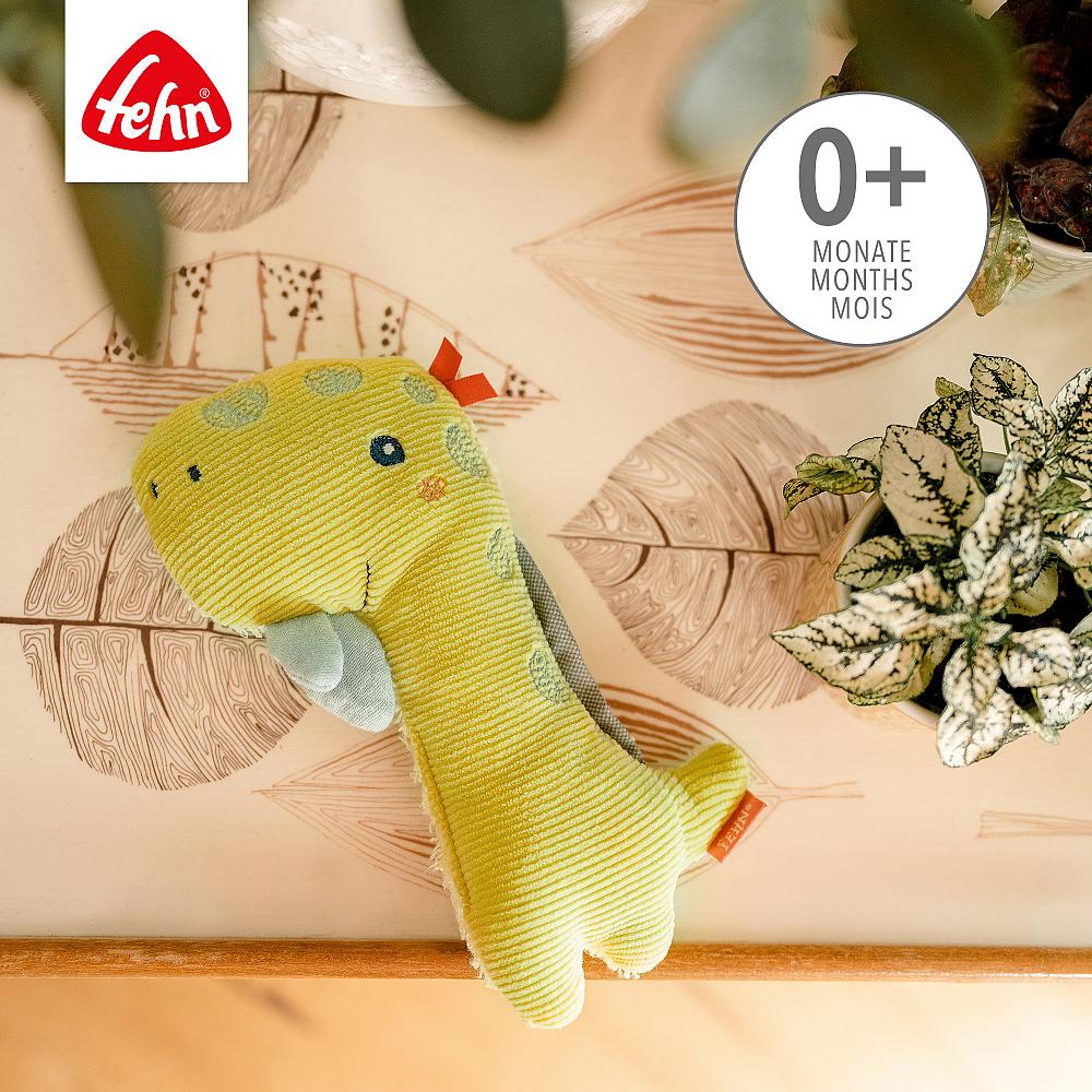 Chocalho dinossauro Fehn amarelo sobre um fundo decorativo, indicando ser adequado para bebés a partir dos 0 meses.