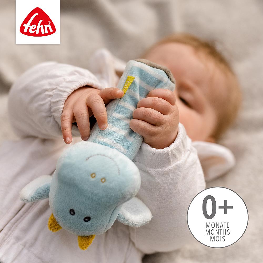 Mãos de bebé a segurar firmemente o Guizo Dragão Fehn, com indicação de idade 0+ meses, promovendo a preensão e o tato.