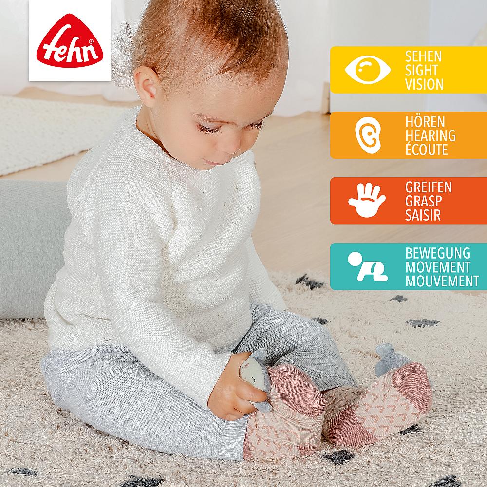 Bebé a interagir com a meia de estimulação Fehn Sereia, com ícones a ilustrar os benefícios: visão, audição, preensão e movimento.