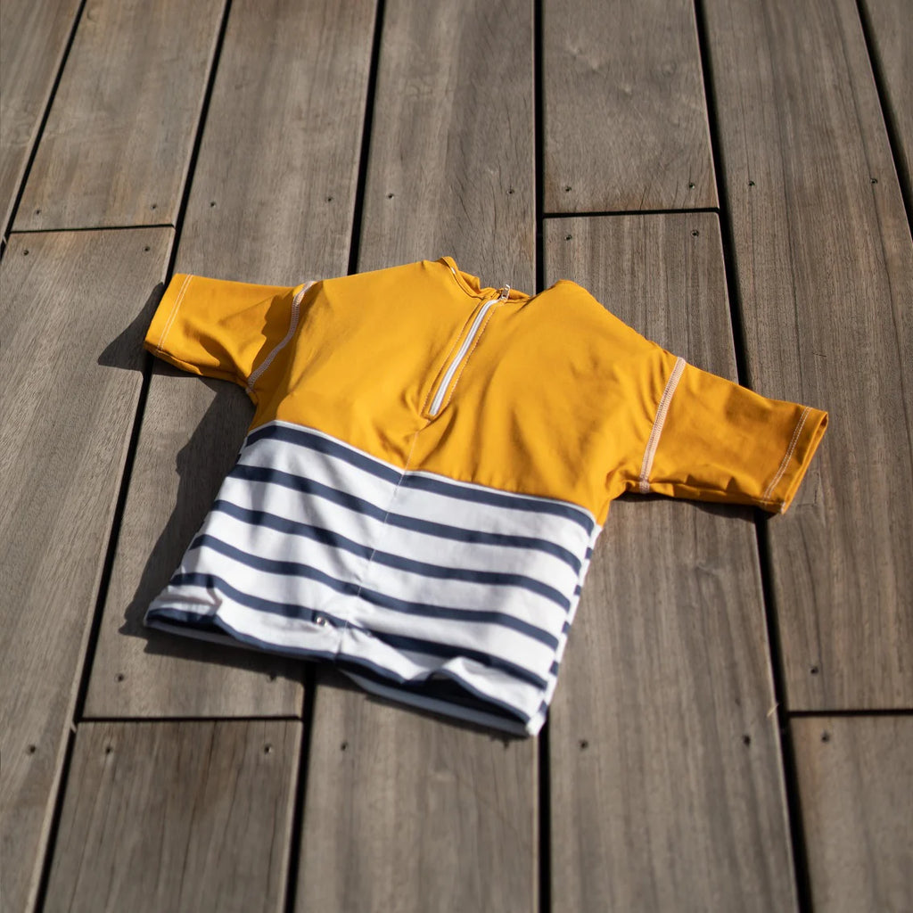 T-shirt anti afogamento Floatee amarela e azul às riscas, com fecho, pousada sobre um deck de madeira ao sol.