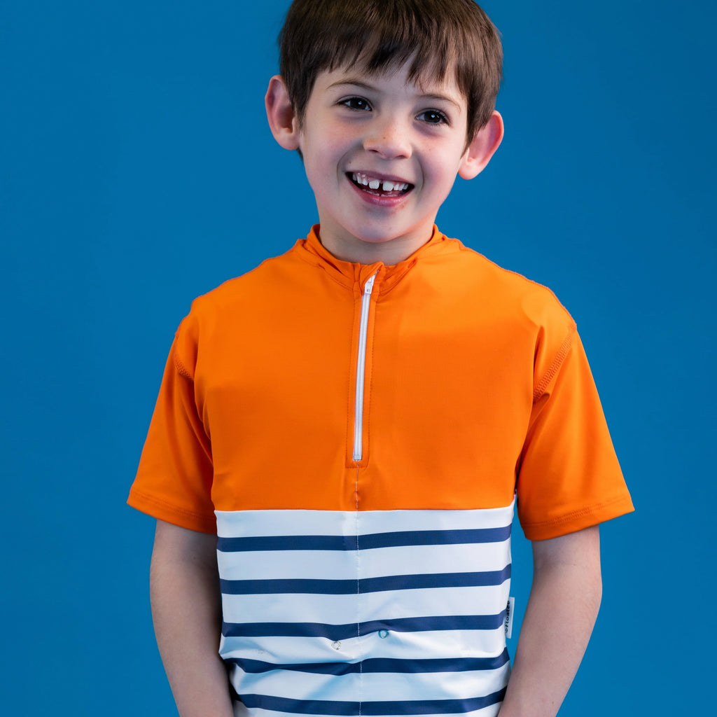 Close-up de um rapaz a sorrir para a câmara, vestindo a T-shirt anti afogamento Floatee laranja e azul às riscas, com fundo azul.