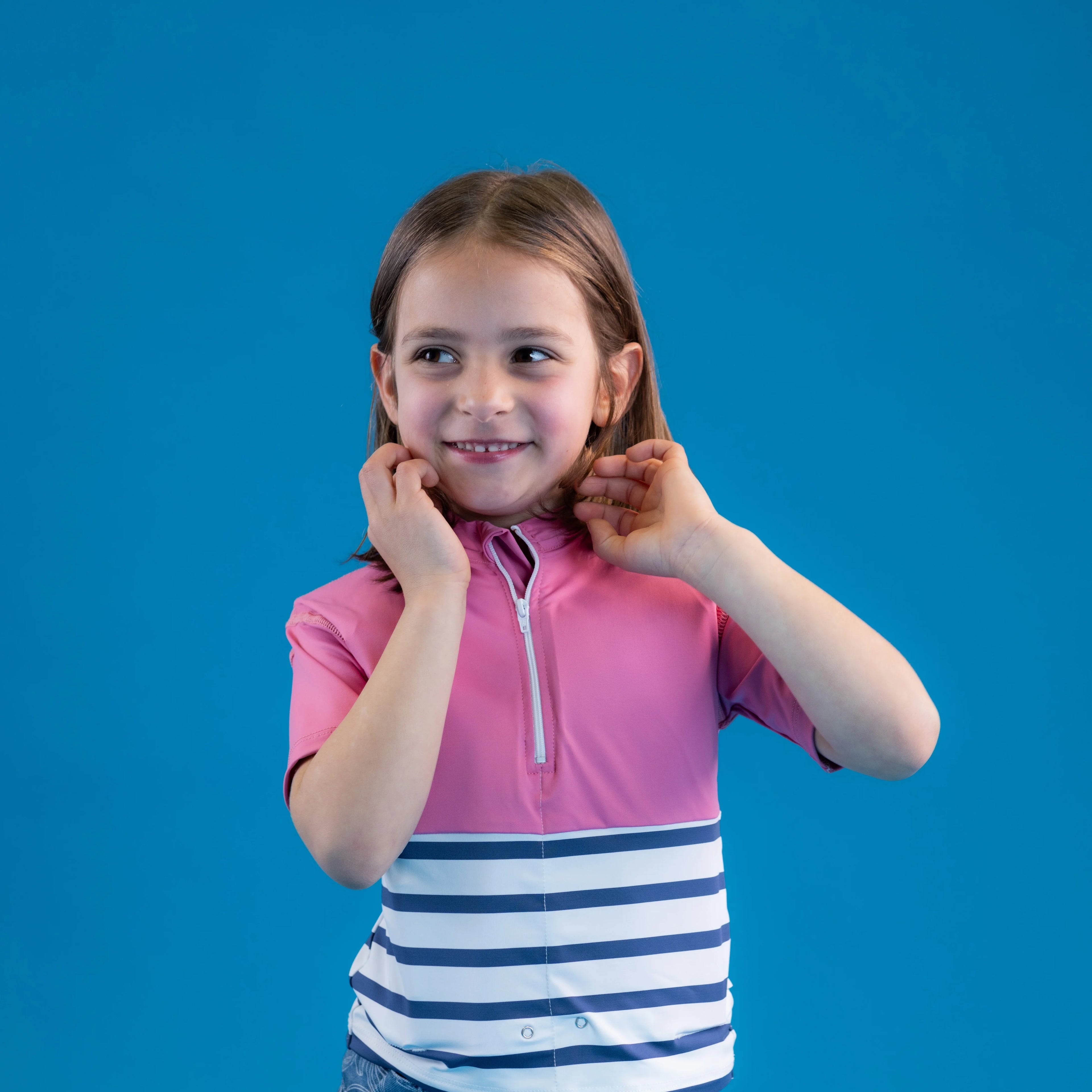 Menina sorridente com a T-shirt anti-afogamento Floatee rosa e branca, ajustando o colarinho, em fundo azul.