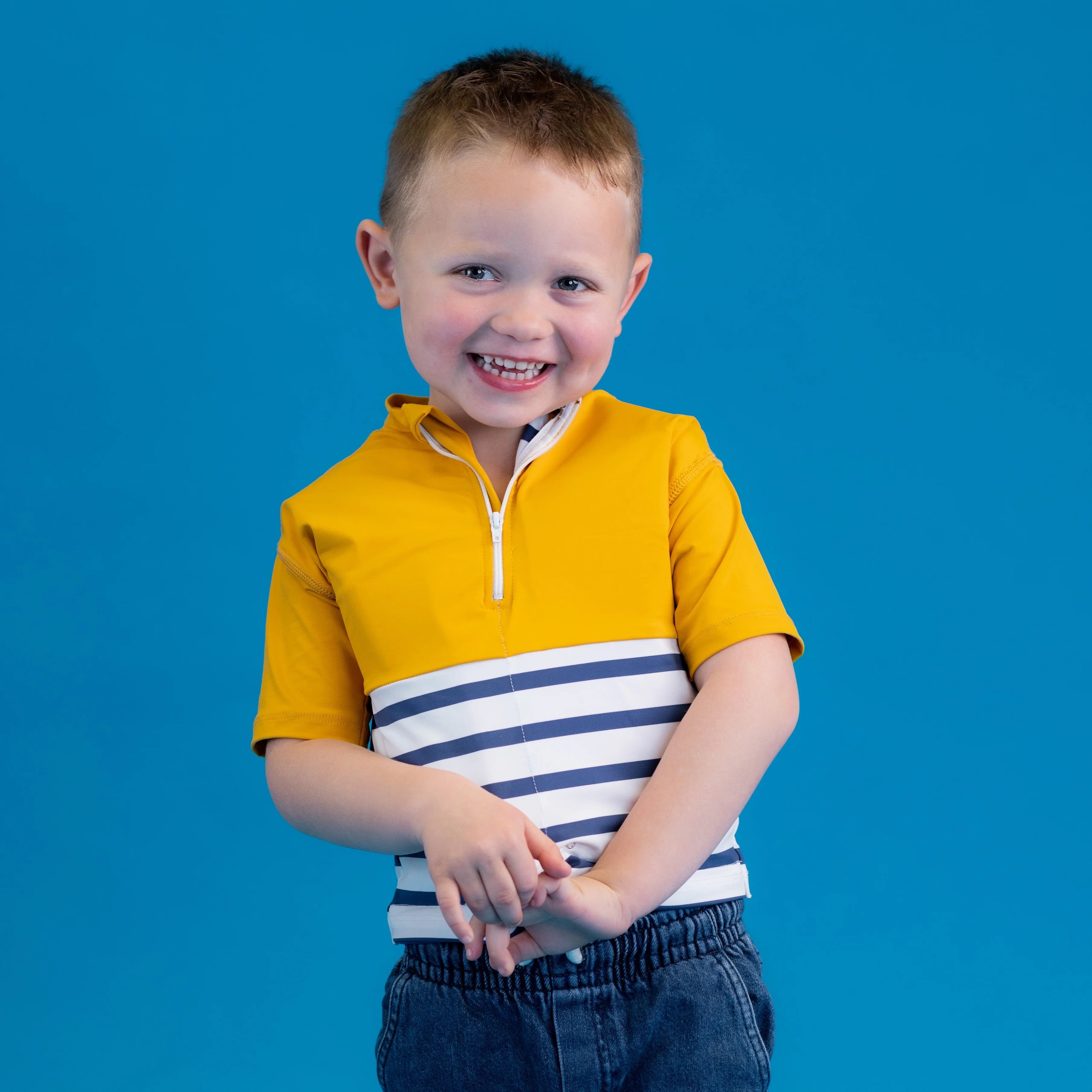 Menino pequeno sorridente com a T-shirt anti-afogamento Floatee amarela e branca, mãos juntas à frente, em fundo azul.
