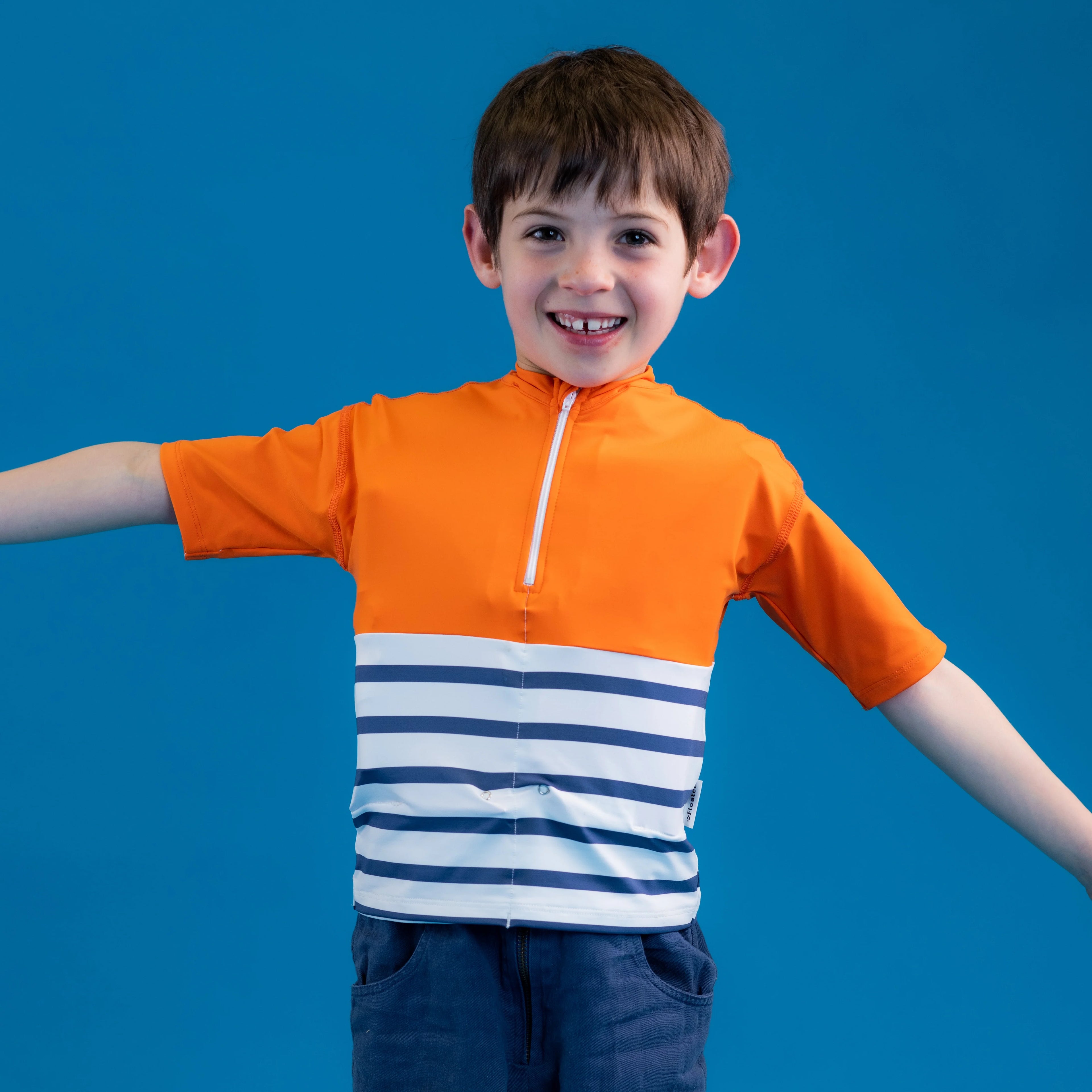 Menino com a T-shirt anti-afogamento Floatee laranja e branca, braços abertos, sorrindo, em fundo azul.