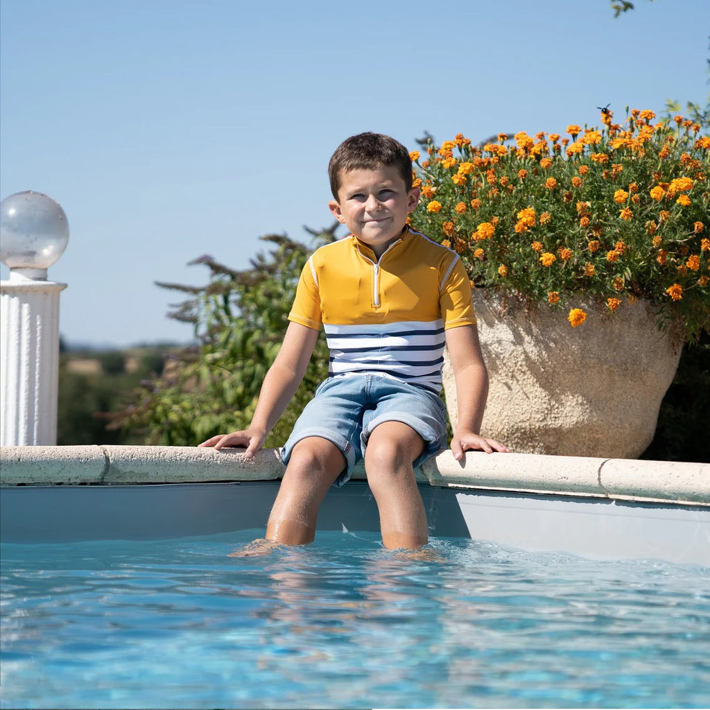 Menino de T-shirt amarela e azul Floatee sentado à beira de uma piscina, com os pés dentro de água, num dia de sol.