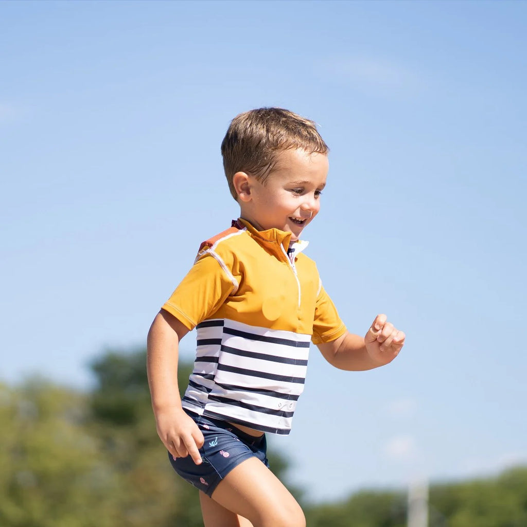 Criança sorridente a correr ao ar livre, vestindo a T-shirt de segurança Floatee amarela e azul às riscas, com céu azul de fundo.