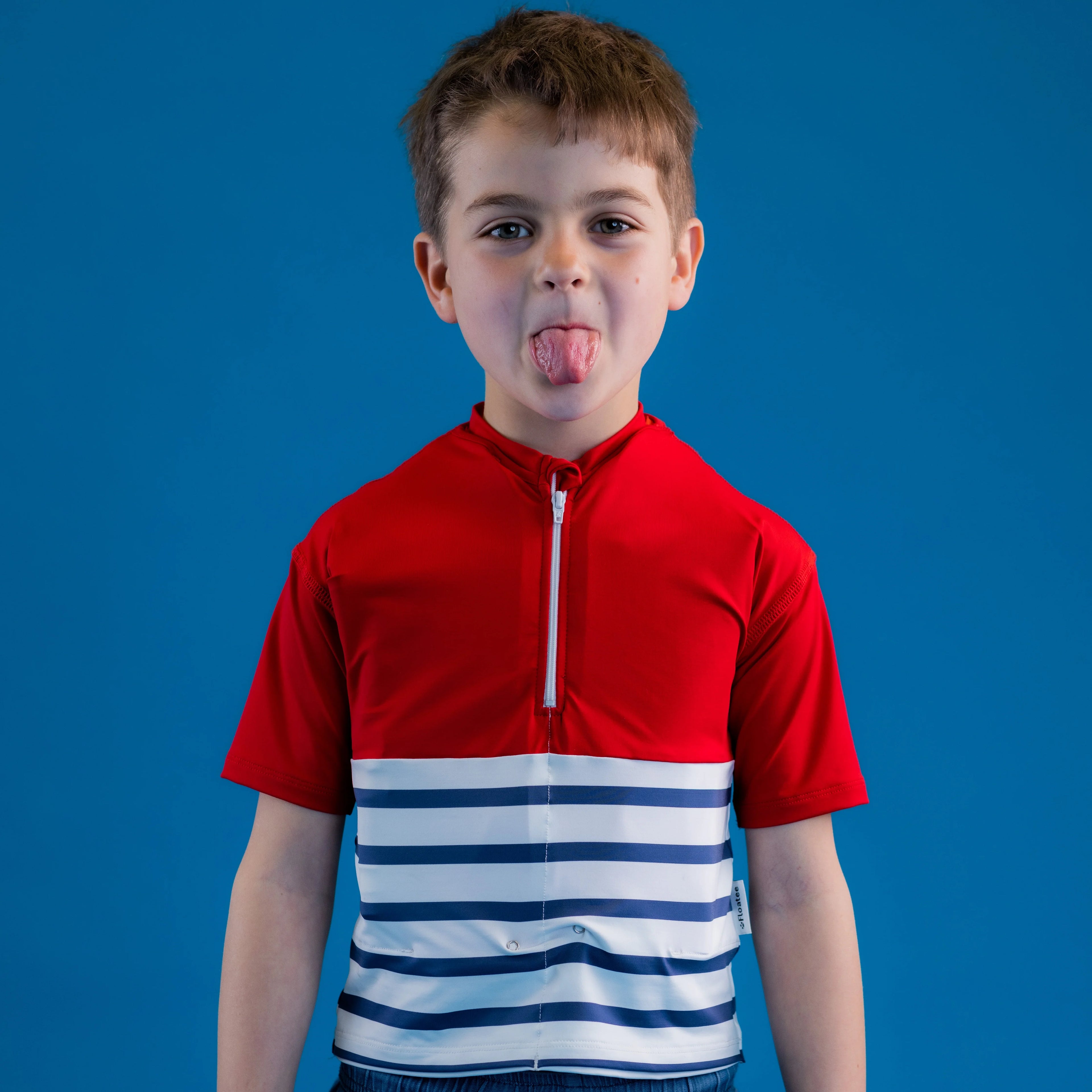 Rapaz a fazer uma careta, a mostrar a língua, vestindo a T-shirt de segurança Floatee vermelha e azul às riscas, com fundo azul.