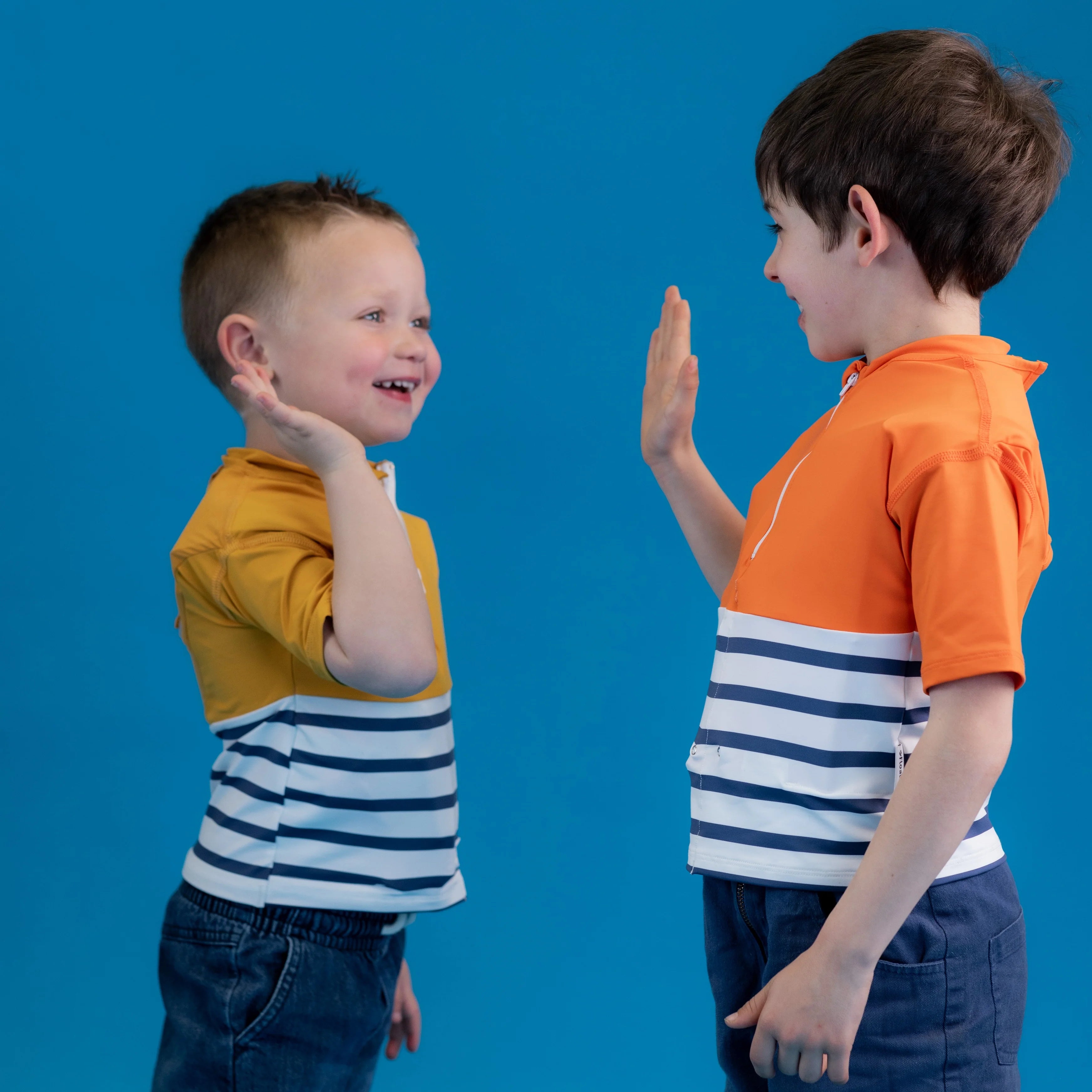 Duas crianças a brincar e a dar as mãos (high-five), uma com a T-shirt Floatee amarela e outra com a versão laranja, em estúdio com fundo azul.